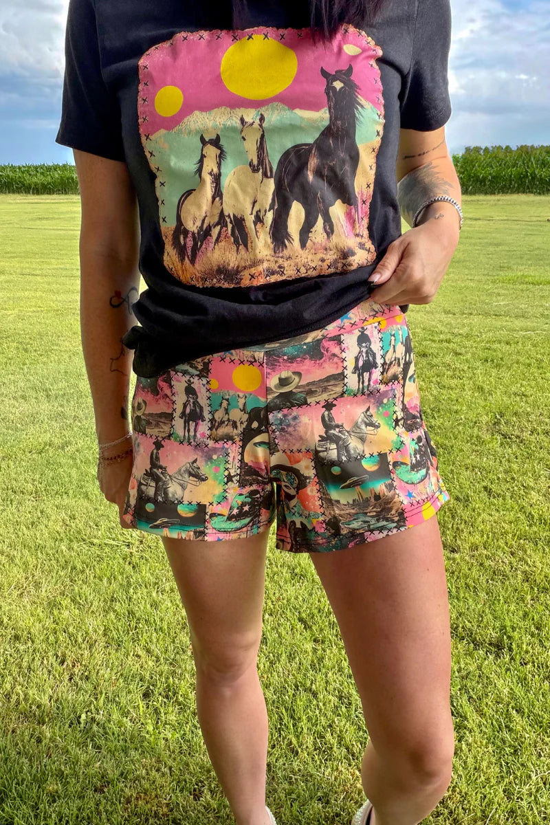 A person standing outdoors on grass wearing a black graphic T-shirt and colorful shorts with a multicolor desert and horse print.