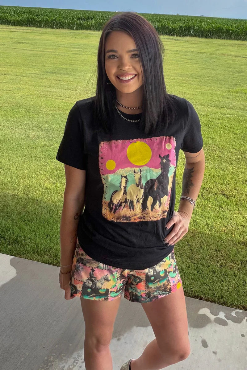 A woman standing outdoors on a concrete walkway with a green grass field behind her, wearing a black graphic t-shirt with a colorful horse print and patterned shorts.