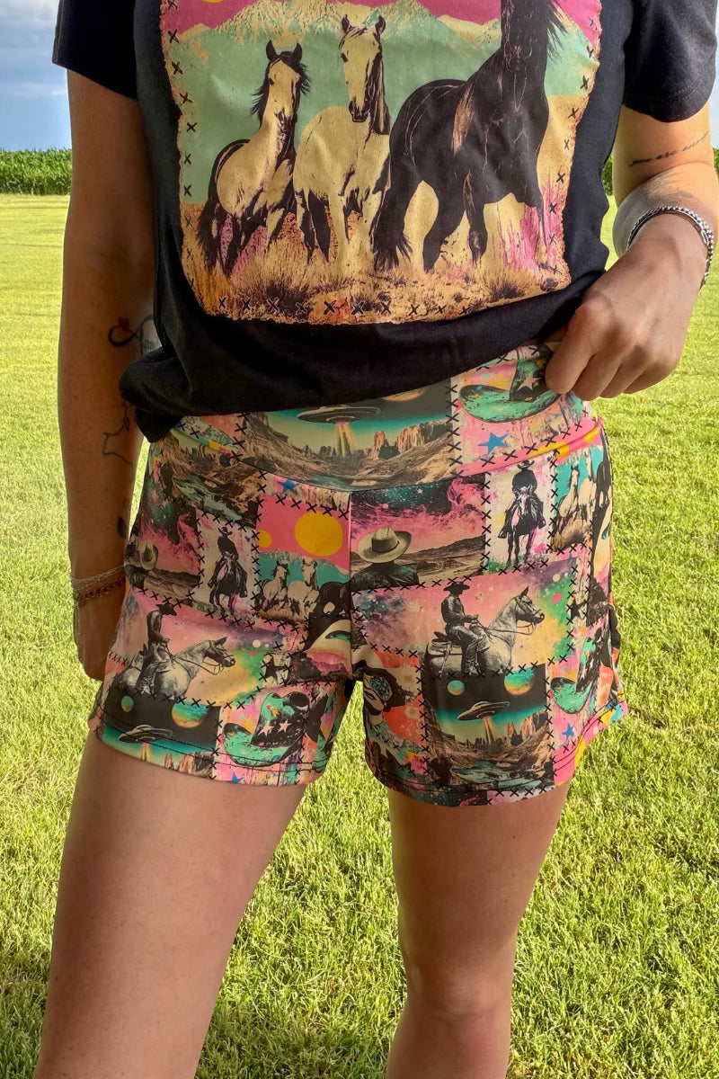 A person outdoors on grass wearing colorful patchwork-print shorts and a black graphic t-shirt with a horse design.